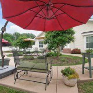 A cozy outdoor patio with a red umbrella, seating, and potted plants on a sunny day.