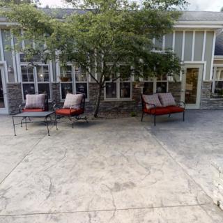A cozy patio area with cushioned chairs under a tree, in front of a building with multiple windows.