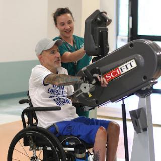 A patient in a wheelchair engages with exercise equipment during a physical therapy session, assisted by a healthcare professional.