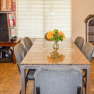 A dining area featuring a long table with gray chairs and a vase of flowers as the centerpiece, complemented by window blinds in the background.