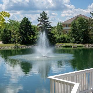 A serene view of a fountain in the center of a small lake, surrounded by trees and a house, with clouds reflecting on the water's surface.