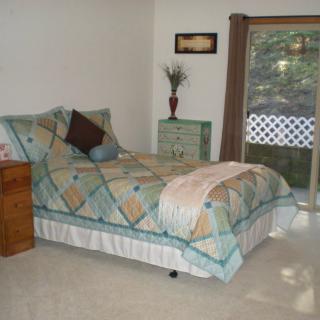 A cozy bedroom with a neatly made bed featuring a colorful quilt, a small dresser with decor, and natural light coming through a sliding glass door.