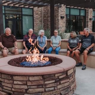 A group of people sit together around a cozy fire pit on a patio.