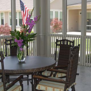 A cozy patio setting with a table, chairs, and a vase of flowers, overlooking an outdoor space with an American flag in the background.