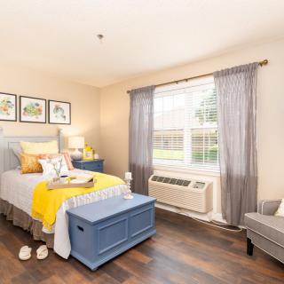 A cozy bedroom with a neatly made bed, decorated in soft colors, featuring a window with curtains and wooden flooring.