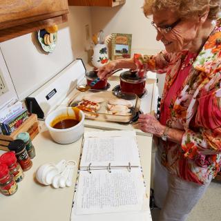 An elderly woman preparing a meal in the kitchen while referencing a recipe book.