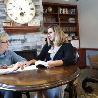 An elderly woman and a younger woman engaged in a discussion over a book in a library setting.