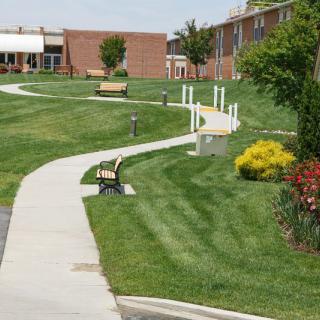 A winding pathway with benches, surrounded by well-maintained grass and landscaping, leading towards buildings.