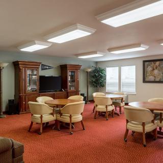 A spacious meeting room with tables and chairs on a red carpeted floor, featuring wooden cabinets and a television.