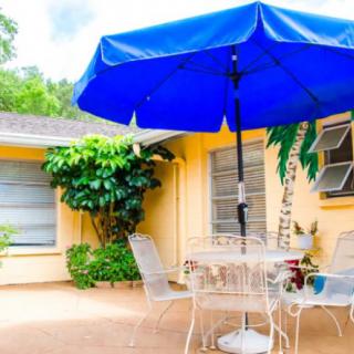 A cozy outdoor patio featuring a blue umbrella over a white table and chairs set, adjacent to a yellow house surrounded by lush plants.