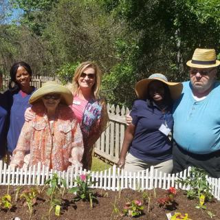 A group of people standing together in a garden with flowers and a white picket fence on a sunny day.