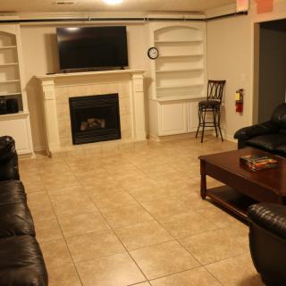 A cozy living room with a television mounted above a fireplace, surrounded by shelves, featuring leather couches and a wooden coffee table on a tiled floor.