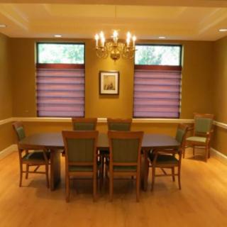 A well-lit dining room with a chandelier, large table, and multiple chairs, framed by two windows.