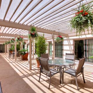 A sunlit patio area with a pergola, featuring a table and chairs surrounded by hanging and potted plants.