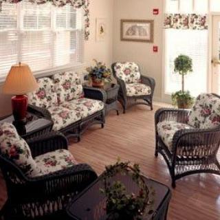 A cozy living room featuring wicker furniture with floral-patterned cushions, a warm lamp, and indoor plants near large windows with blinds.