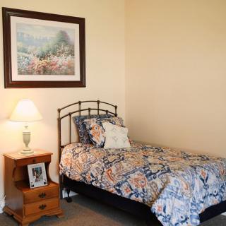 A cozy bedroom with a single bed covered in a floral comforter, a wooden nightstand holding a lamp and framed photo, and a painting on the wall.