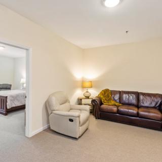 A cozy living room featuring a leather sofa, an armchair, and a lamp placed in the corner. A doorway leads to a bedroom.