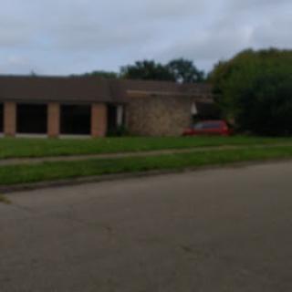 A view of a residential building with a grassy front yard and a red car parked nearby, under a cloudy sky.