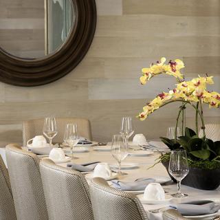 An elegantly set dining table with a yellow orchid centerpiece and wine glasses, surrounded by plush chairs and accentuated by a large round mirror on the wall.
