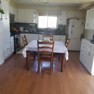 A spacious kitchen with a dining table and wooden chairs, featuring white cabinets and a large window.