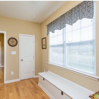 A bright hallway featuring a window with patterned curtains, a bench, and hardwood flooring.