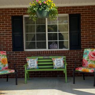 A cozy patio setup with a green bench, floral cushioned chairs, and a hanging plant in front of a brick wall.