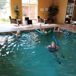 A group of seniors participating in water aerobics at an indoor pool, using pool noodles for support.