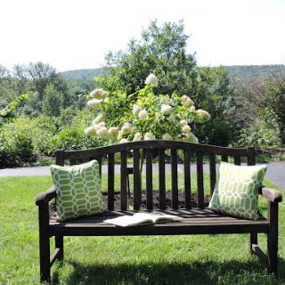 A serene garden scene featuring a wooden bench adorned with decorative pillows, surrounded by lush greenery and blooming flowers.