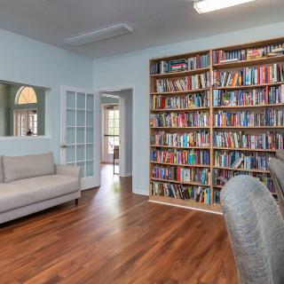 A cozy library room with a large bookshelf filled with books, featuring a sofa and chairs on a hardwood floor.