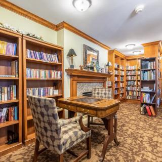 A cozy library with wooden bookshelves, a checkered chair, and a central wooden table in front of a fireplace.