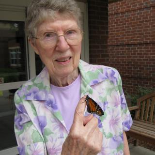 An elderly woman smiling while holding a butterfly on her hand.