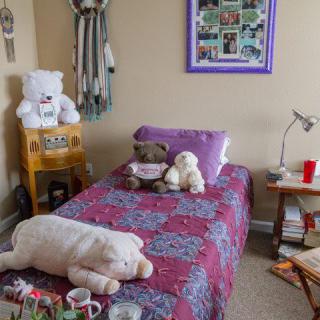 A cozy bedroom with a single bed adorned with colorful bedding and stuffed animals, surrounded by shelves, books, and a lamp.