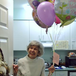 An elderly woman smiles joyfully while holding a bunch of colorful balloons in a kitchen setting.
