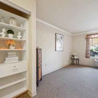 A cozy living room with a built-in shelf and carpet flooring, featuring natural light from a window with checkered curtains.