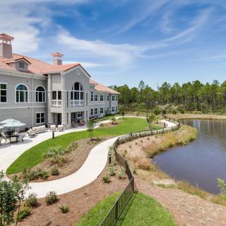 A large, elegant building with a red roof by a scenic lake, surrounded by landscaped pathways and greenery under a blue sky.