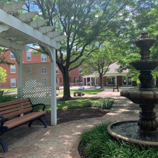 A serene garden scene featuring a bench, fountain, and gazebo under lush trees.