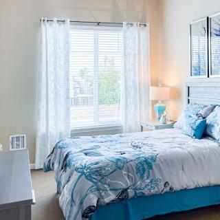 A cozy bedroom with blue and white decor, featuring a bed by the window with sheer curtains.
