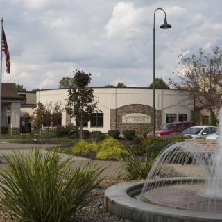 A building with a fountain in the foreground, accompanied by a flag and a parking area under a cloudy sky.