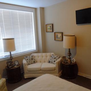 A cozy bedroom with classic decor, featuring a seating area with lamps and a window with blinds.