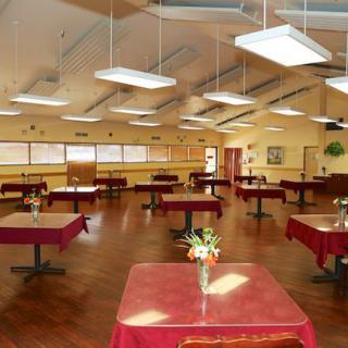 A spacious dining hall with neatly arranged tables and chairs, featuring a wooden floor and ambient lighting.
