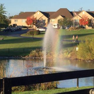 A tranquil park scene featuring a fountain creating a rainbow over a pond, with autumn trees and houses in the background.