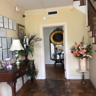 A well-decorated hallway featuring a table with a lamp and plant, framed certificates on the wall, and a staircase leading upwards. A large mirror is visible through the doorway along with colorful floral arrangements.