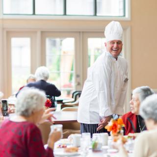 A chef interacts with seniors in a dining area, creating a welcoming atmosphere.