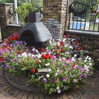 A vibrant garden display with colorful flowers surrounding a chiminea against a brick wall backdrop.