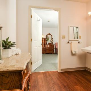 A view of a bathroom with wooden flooring, featuring a sink, mirror, towel rack, and plant, leading into a bedroom with furniture.