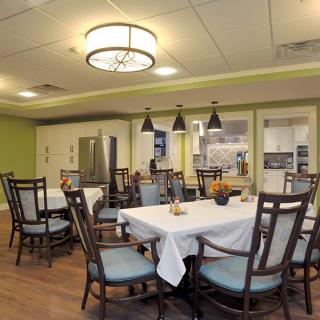A well-lit dining room with neatly arranged tables and chairs, adjacent to a kitchen area.