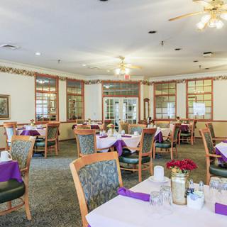 A well-lit dining area with neatly arranged tables and chairs, adorned with purple napkins and simple floral centerpieces.