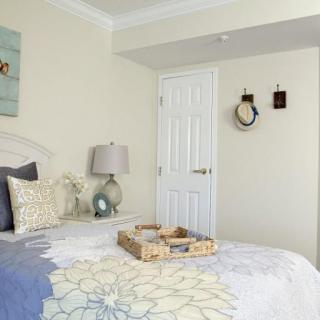 A cozy bedroom with light decor, featuring a bed with floral bedding, a bedside lamp, and wall decorations including artwork and a clock.