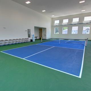 An indoor sports facility featuring a blue tennis court with a green floor, surrounded by windows and lined with chairs.