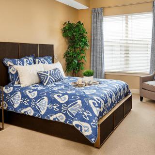A cozy bedroom with a patterned blue bedspread, large window, and decorative plant.